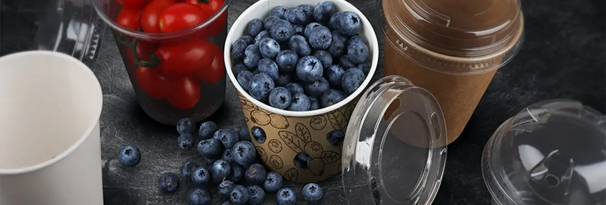 Auf einem Tisch stehen mehrere Becher aus Karton und Kunststoff mit durchsichtigen Kunststoffdeckel, gefüllt mit Blaubeeren oder Tomaten