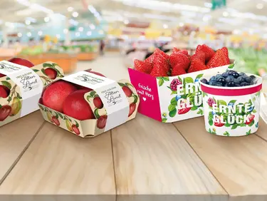 Auf einem Tisch in einem Einkaufsmarkt stehen verschiedene Obstverpackungen mit Äpfeln, Erdbeeren und Blaubeeren.